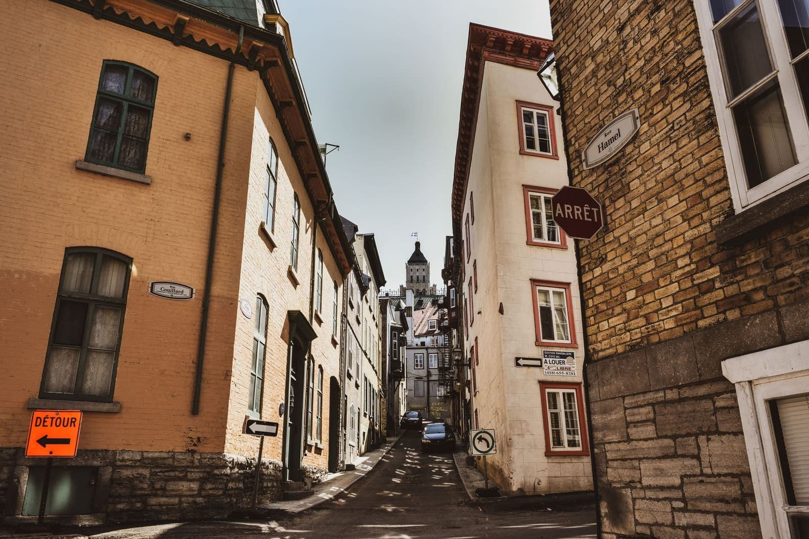 A narrow street in late afternoon shade, a bakery light at one end.